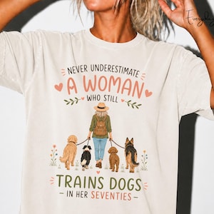 Könnte beinhalten: Ein cremefarbenes T-Shirt mit einer grafischen Illustration einer Person mit Hut und Rucksack, die vier Hunde an der Leine führt. Der Text lautet: NEVER UNDERESTIMATE A WOMAN WHO STILL TRAINS DOGS IN HER SEVENTIES, umrahmt von floralen Akzenten.