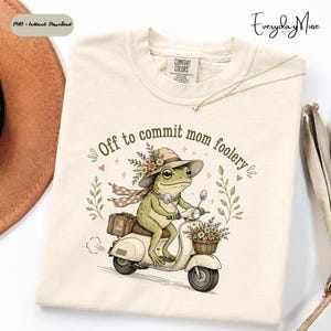 May include: Cream-colored t-shirt featuring a whimsical illustration of a frog riding a scooter. The frog wears a hat and scarf, with the text "Off to commit mom foolery." The scooter has a basket of flowers. A necklace is draped on the shirt.