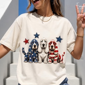 May include: A cream-coloured short-sleeve t-shirt featuring a graphic of three Basset Hound dogs. The left dog has a blue star-patterned coat, the centre dog is white, and the right dog has red and white stripes. Two blue stars and one red star are above.