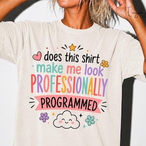 May include: A beige short-sleeve t-shirt featuring the text does this shirt make me look PROFESSIONALLY PROGRAMMED in colourful lettering. The design includes a pink heart, a yellow star, a smiling cloud, and small flowers in orange, purple, and teal.