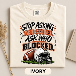 May include: An ivory t-shirt featuring a graphic with the text "STOP ASKING WHO SCORED. ASK WHO BLOCKED." in black and orange lettering. Below the text is an illustration of a brown football and a white football helmet resting on green grass.