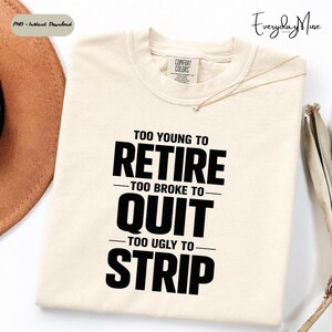 May include: Cream-colored t-shirt with the text "TOO YOUNG TO RETIRE - TOO BROKE TO QUIT - TOO UGLY TO STRIP" in bold black letters. The shirt has a Comfort Colors tag and a gold necklace.