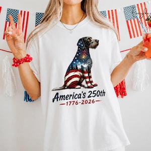 Puede incluir: Una camiseta blanca con el gráfico de un perro cuyo pelaje tiene el diseño de la bandera estadounidense, incluyendo estrellas, rayas y un paisaje montañoso. Debajo del perro, el texto dice America's 250th 1776-2026 en letras azul y rojo.