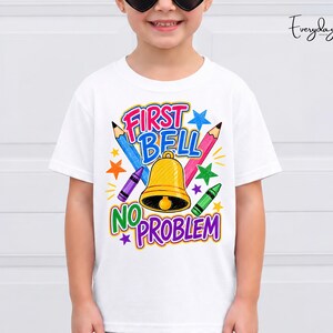 以下が含まれることがあります： FIRST BELL NO PROBLEMというテキストが書かれたカラフルなグラフィックが特徴の白いTシャツ。デザインには、黄色いベル、青とピンクの鉛筆、紫と緑のクレヨン、そして中央のテキストを囲む様々なマルチカラーの星が含まれています。
