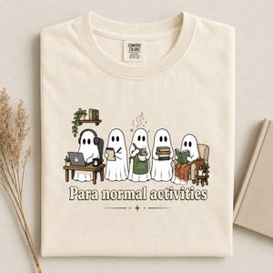 Peut inclure: Un t-shirt couleur crème avec un motif représentant cinq fantômes blancs occupés à diverses activités, notamment travailler sur un ordinateur portable, écrire, cuisiner, tenir des livres et lire. Le texte "Para normal activities" est imprimé en dessous.
