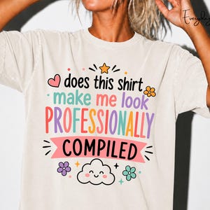 Op de afbeelding: Een beige t-shirt met korte mouwen en de tekst "does this shirt make me look PROFESSIONALLY COMPILED" in kleurrijke letters. Het ontwerp bevat een kleine oranje ster, een roze hartje, een bloem en een lachende wolk onderaan de afbeelding.