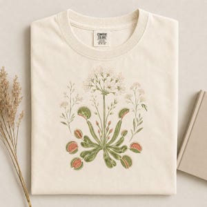 May include: A cream-coloured t-shirt featuring a botanical illustration of a Venus flytrap plant with green leaves, reddish-pink traps, and white flowers. The shirt has a crew neckline and a Comfort Colors brand label printed on the inside collar.