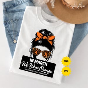 Op de afbeelding: Wit T-shirt met een afbeelding van een vrouw met een oranje strik, zonnebril met oranje lintontwerpen en de tekst "In March We Wear Orange" voor Multiple Sclerose Awareness. Het shirt is op een blauwe spijkerbroek gevouwen.