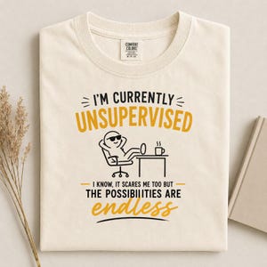 Op de afbeelding: Een crèmekleurig t-shirt met een afbeelding van een stokfiguurtje met een zonnebril, zittend in een stoel met de voeten op een bureau naast een koffiemok. De tekst luidt: I'M CURRENTLY UNSUPERVISED. I KNOW, IT SCARES ME TOO BUT THE POSSIBILITIES ARE ENDLESS.