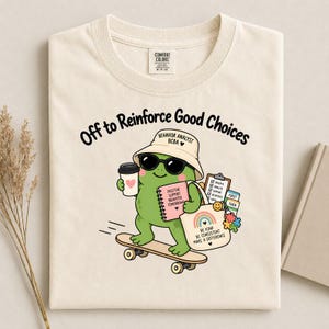 Könnte beinhalten: Ein cremefarbenes T-Shirt mit einer Grafik eines grünen Frosches, der einen Fischerhut und eine Sonnenbrille trägt und auf einem Skateboard fährt. Der Frosch hält einen Kaffeebecher und eine Tasche. Text: Off to Reinforce Good Choices, Behavior Analyst BCBA.