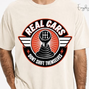 Puede incluir: Una camiseta de color crema con un gráfico de una palanca de cambios manual negra. El diseño incluye el texto "REAL CARS" y "DONT SHIFT THEMSELVES" en blanco, sobre un fondo circular naranja y negro con alas a los lados.