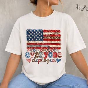 Könnte beinhalten: Cremefarbenes T-Shirt mit einem verwaschenen Design der amerikanischen Flagge und Leopardenmuster. Der Text "Remember Everyone Deployed" ist in verschiedenen Schriftarten und Mustern dargestellt. Das T-Shirt hat einen lässigen, patriotischen Stil.