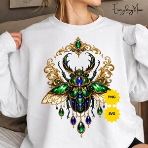 May include: White sweatshirt featuring a detailed illustration of a jeweled beetle in shades of green, blue, and gold. The design includes ornate filigree and dangling gemstone accents. Two yellow starbursts with "PNG" and "SVG" text are also visible.