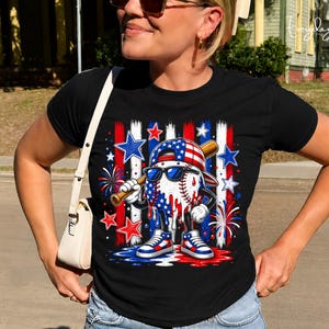 May include: A black t-shirt featuring a graphic of a baseball character wearing a flag-patterned cap and sunglasses, holding a wooden bat. The design includes red, white, and blue stars, vertical stripes, and fireworks, with a melting paint effect.