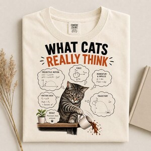 Op de afbeelding: Een crèmekleurig t-shirt met een afbeelding van een cyperse kat die een mok van een tafel stoot. De tekst luidt WHAT CATS REALLY THINK, omringd door natuurkundige vergelijkingen voor projectielbeweging, kracht, impuls, wrijving en traject.