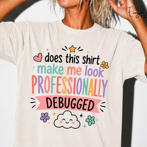 May include: A beige t-shirt featuring a colorful graphic with the text "does this shirt make me look PROFESSIONALLY DEBUGGED". The design includes a small orange star, a pink heart, a smiling cloud, and several small flowers in purple and teal.