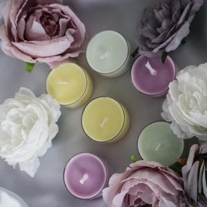 Könnte beinhalten: Draufsicht auf sieben Teelichter in Gelb, Grün und Lila, umgeben von weißen, lila und grauen Rosen. Die Kerzen sind auf einer silbernen Satin-Oberfläche angeordnet und erzeugen eine weiche, romantische Atmosphäre.