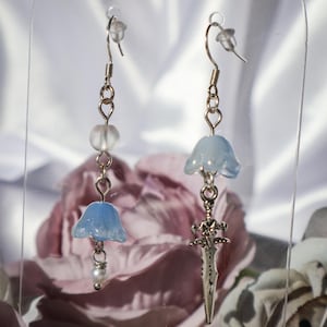 May include: A pair of dangle earrings with a whimsical design. One earring features a light blue, bell-shaped bead, a clear bead, and a small pearl. The other earring has a light blue bell-shaped bead and a silver sword charm.