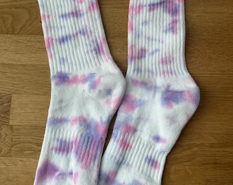 May include: A pair of white crew socks with a tie-dye pattern in pink and purple hues. Each sock features a black Nike swoosh logo near the top.