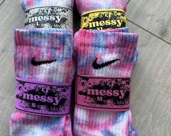 May include: Four pairs of tie-dye socks with a white Nike swoosh logo. The socks are in different sizes: S, M, L, and XL. The socks are packaged with a black and white label that says "messy" and the size.