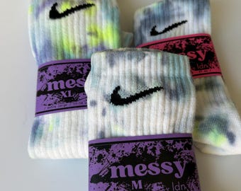 May include: Three pairs of tie-dye socks with a black Nike swoosh logo. The socks are in different color combinations of blue, green, and white. The socks are labeled with the brand "messy Idn" and the size "M" for medium, "XL" for extra large, and "S" for small.
