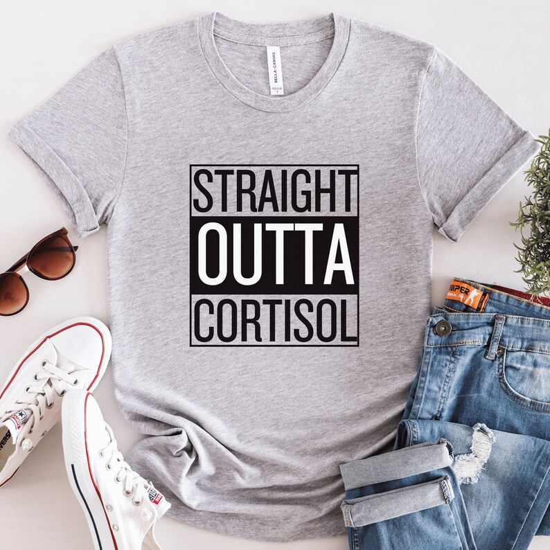 May include: A light gray t-shirt with the text "STRAIGHT OUTTA CORTISOL" in a black and white design. The shirt is displayed with sunglasses, white sneakers, and blue jeans.