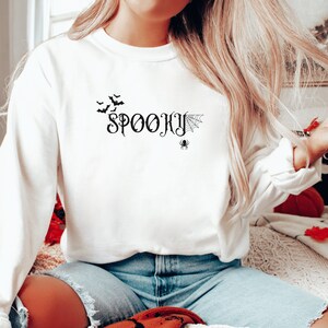 May include: White long-sleeve sweatshirt with the text "SPOOKY" in black script font. The design also includes a spider web and a spider in black.