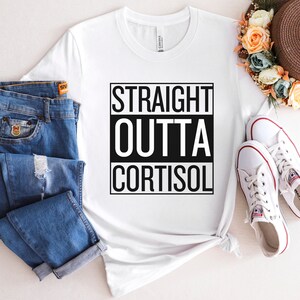 May include: White t-shirt with the text "STRAIGHT OUTTA CORTISOL" in a black box. The shirt is paired with blue jeans and white sneakers. A straw hat with floral accents is also visible.