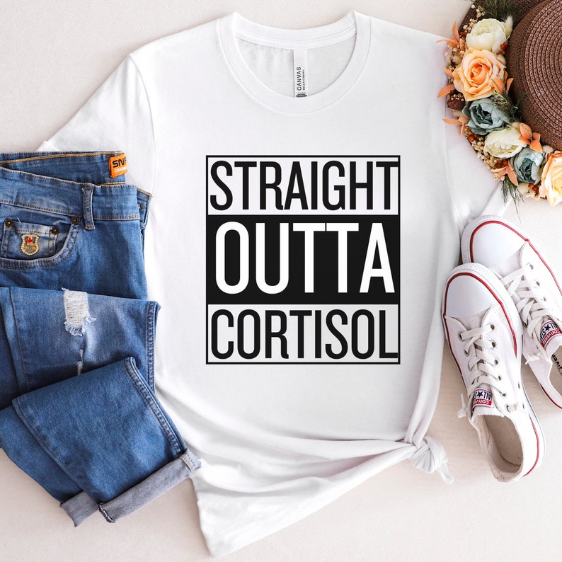 May include: White t-shirt with the text "STRAIGHT OUTTA CORTISOL" in a black box. The shirt is paired with blue jeans and white sneakers. A straw hat with floral accents is also visible.