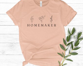 May include: A peach-colored t-shirt with a simple design of three stylized flowers and the word "HOMEMAKER" printed in black.