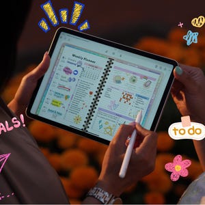 May include: A person holding a tablet displaying a weekly planner with colorful graphics and text. The planner includes sections for grocery shopping, goals, and to-do lists. The word "GOALS!" is written in pink, and a pink paper airplane is drawn on the image.