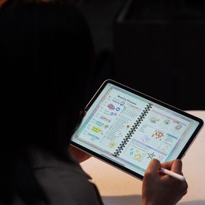 May include: A person holding a tablet displaying a colorful weekly planner. The planner features a spiral binding and is filled with illustrations and text. A stylus is used to interact with the planner. The background is dark.