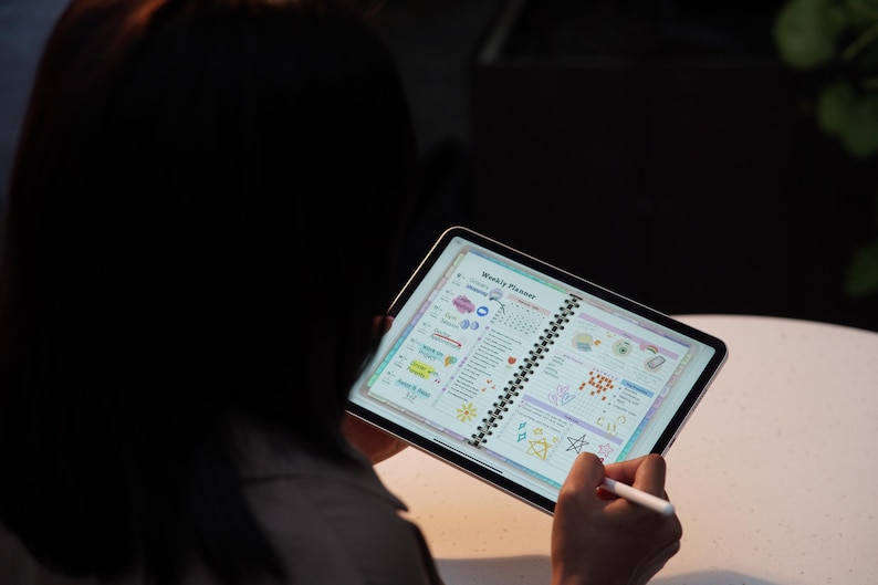 May include: A person holding a tablet displaying a colorful weekly planner. The planner features a spiral binding and is filled with illustrations and text. A stylus is used to interact with the planner. The background is dark.