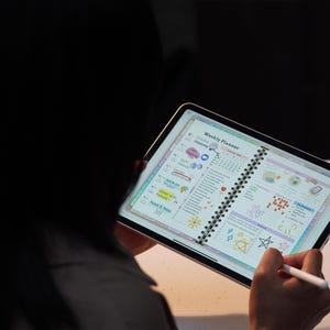 May include: A digital tablet displaying a weekly planner with colorful illustrations and handwritten notes. The tablet is held by a person, with a stylus in hand. The planner features sections for appointments, to-do lists, and notes.