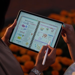 May include: A digital weekly planner displayed on a tablet screen, featuring colorful icons and handwritten notes. The tablet is held by two people, one using a stylus to write. The background is blurred with orange flowers.