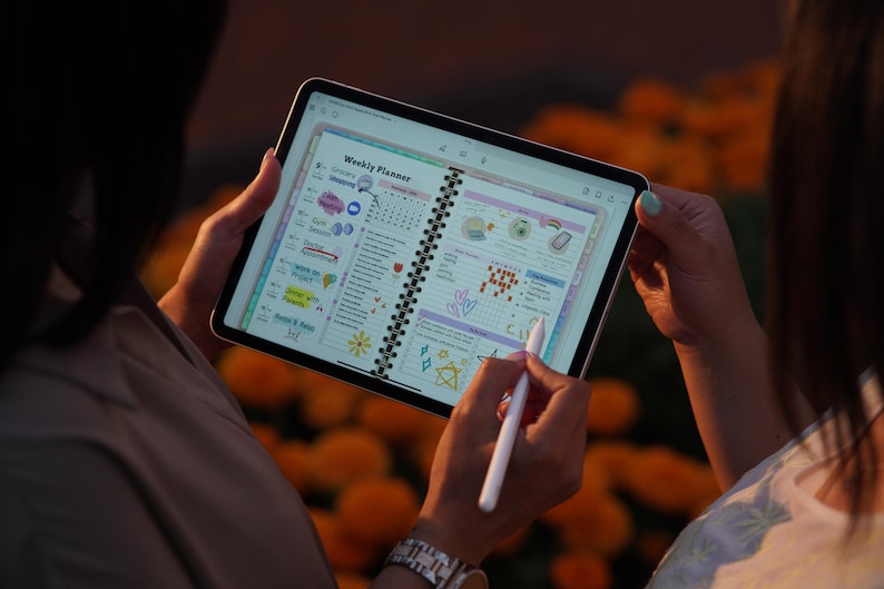 May include: A digital weekly planner displayed on a tablet screen, featuring colorful icons and handwritten notes. The tablet is held by two people, one using a stylus to write. The background is blurred with orange flowers.