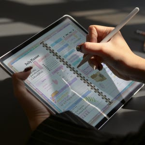May include: A digital daily planner displayed on a tablet, held by a person. The planner is open to a page with colorful sections and the text "Daily Planner." A white stylus is used to write on the screen. The tablet has a silver frame.