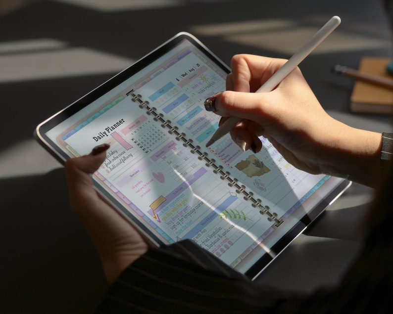 May include: A digital daily planner displayed on a tablet, held by a person. The planner is open to a page with colorful sections and the text "Daily Planner." A white stylus is used to write on the screen. The tablet has a silver frame.