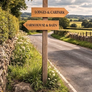 May include: A wooden signpost with two signs pointing in opposite directions. The top sign reads "Lodges & Carpark" and the bottom sign reads "Farm House & Barn". The sign is set on a country road with a stone wall and green fields.