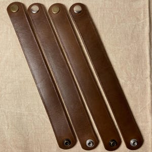 May include: Four brown leather straps with metal snap closures. Each strap has a different color metal snap, including brass and silver. The straps are arranged diagonally on a beige fabric surface.