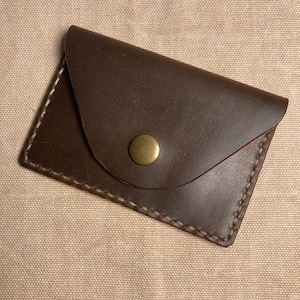 May include: A small, brown leather card holder with a snap closure. The rectangular card holder has rounded edges and a flap secured by a brass-coloured snap. The edges are stitched with a light-coloured thread.