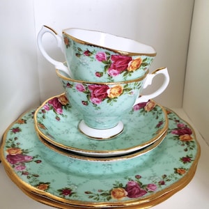 May include: A stack of two porcelain teacups, a saucer, and two plates featuring a light mint green background with a floral pattern of pink and yellow roses. Each piece is finished with a gold-trimmed scalloped edge and white interior.