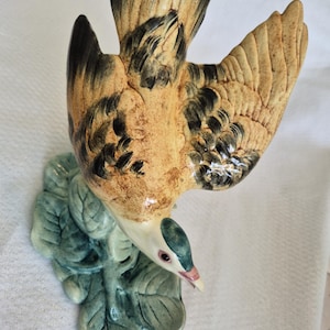 May include: A ceramic bird figurine featuring a tan and brown body with dark wing tips, perched atop a textured green leaf base. The bird is depicted in a downward-facing position with its wings partially spread, showcasing detailed feather textures.