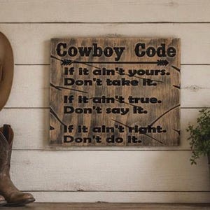 May include: A rectangular wooden sign with a distressed finish featuring the black text Cowboy Code, If it ain't yours. Don't take it. If it ain't true. Don't say it. If it ain't right. Don't do it. The sign is decorated with a black arrow graphic.