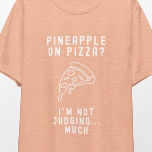 May include: A peach-colored t-shirt with the text "Pineapple on pizza? I'm not judging... much" in white. A white outline of a pizza slice is above the text.
