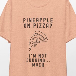 May include: A peach-colored t-shirt with a black graphic of a pizza slice with the text "Pineapple on pizza? I'm not judging... much".