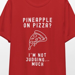 May include: Red t-shirt with white text that reads "Pineapple on pizza? I'm not judging... much." and a white outline of a pizza slice with pineapple on top.