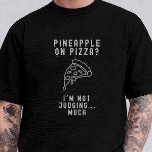 May include: A black t-shirt with a white graphic of a pizza slice and the text "Pineapple on pizza? I'm not judging... much".