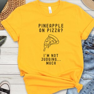 May include: Yellow t-shirt with a black graphic of a pizza slice with pineapple on top. The text reads "Pineapple on Pizza? I'm not judging... much."