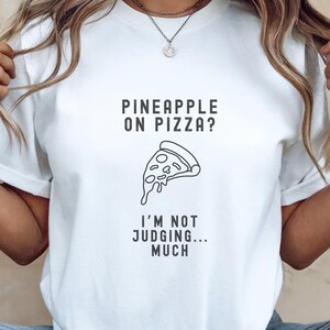 May include: White t-shirt with a black graphic that says "Pineapple on pizza? I'm not judging... much". The graphic also includes a slice of pizza with a dripping cheese.